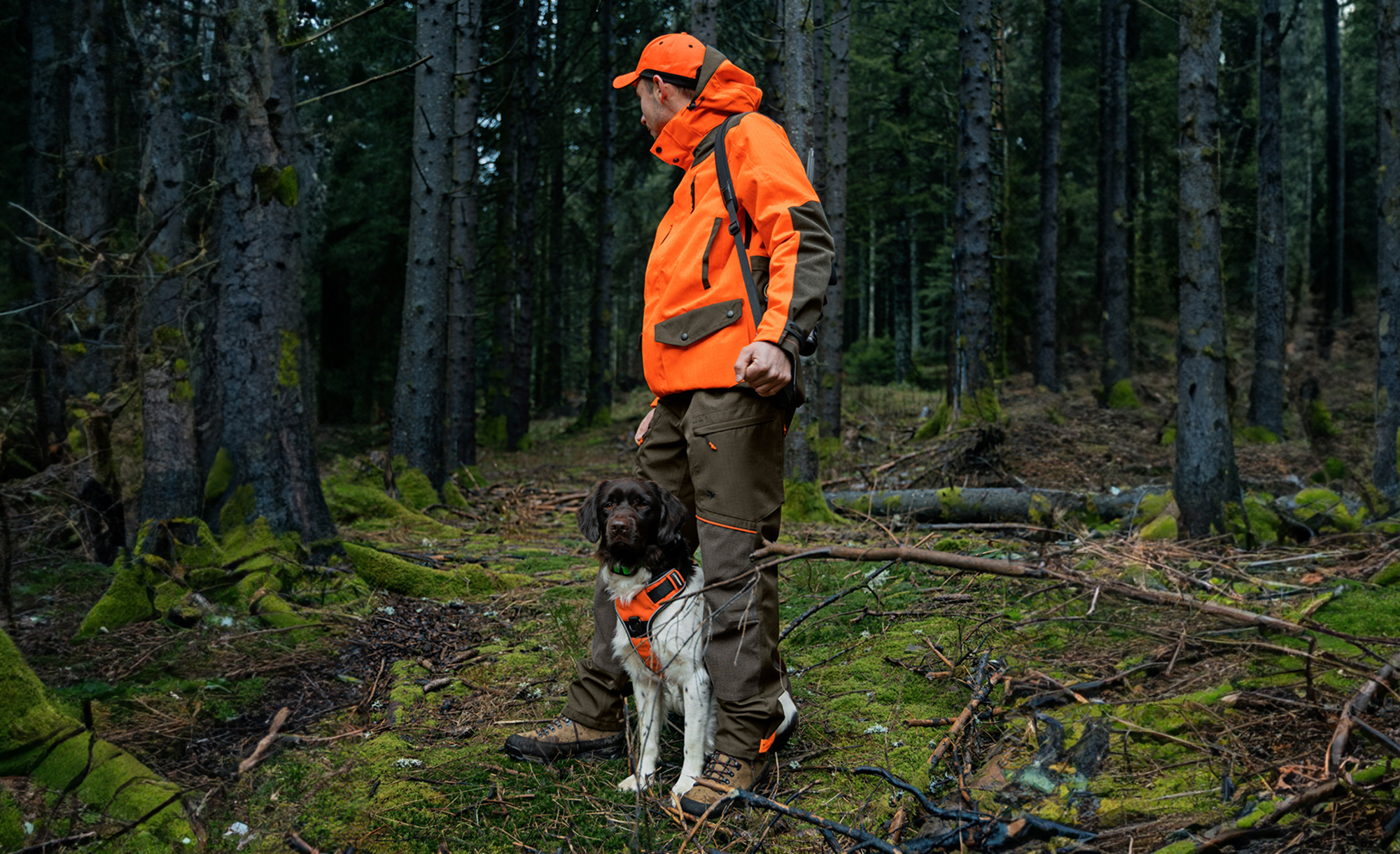 Een jager in oranje en bruine kleding staat met zijn hond in een met mos bedekte bosomgeving. Beiden dragen opvallende signaalvesten voor betere zichtbaarheid.