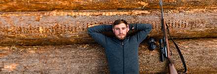 Een man ligt ontspannen met de armen achter het hoofd op gestapelde boomstammen; naast hem ligt een jachtgeweer met richtkijker.