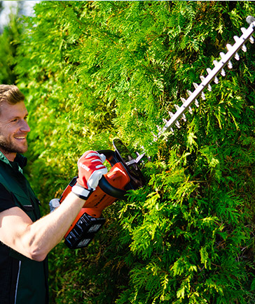 Man in groene werkkleding snoeit een dichte groene haag met een elektrische heggenschaar.