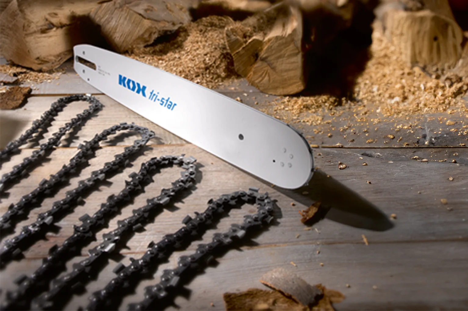 Een motorzaaggeleiderail met het opschrift „KOX tri-star” ligt op een houten oppervlak. Daarnaast liggen meerdere zaagkettingen netjes uitgestald, met op de achtergrond houtstukken en zaagsel.