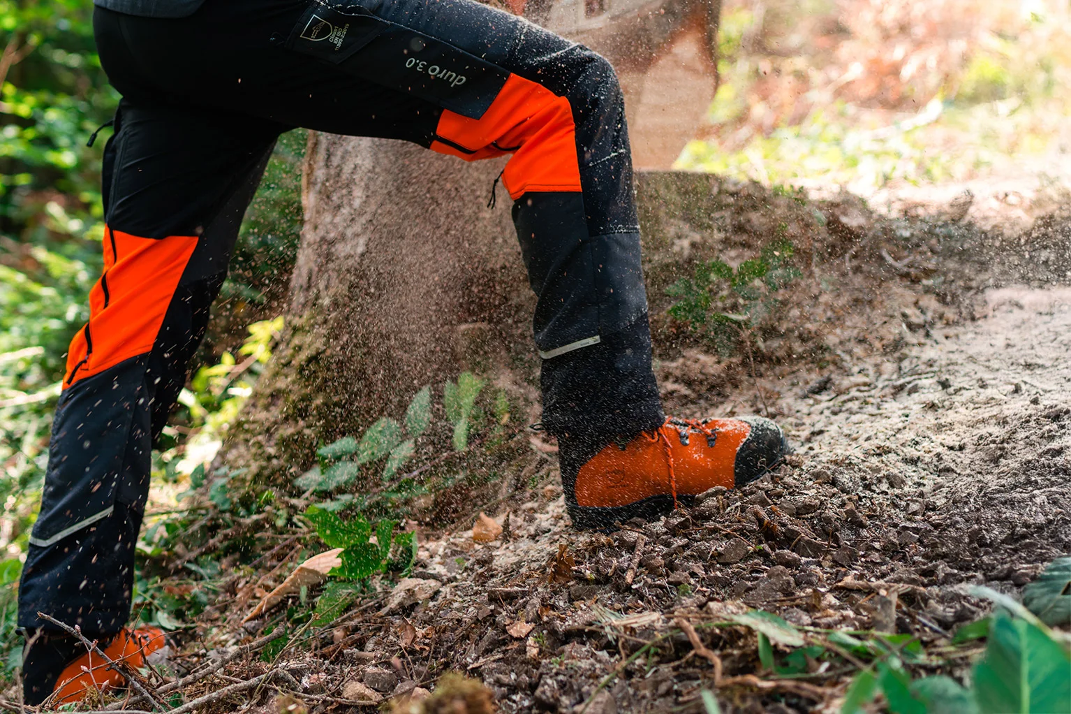 Benen van een persoon met een beschermende zaagbroek en veiligheidsschoenen tijdens boswerkzaamheden, houtspaanders vliegen rond