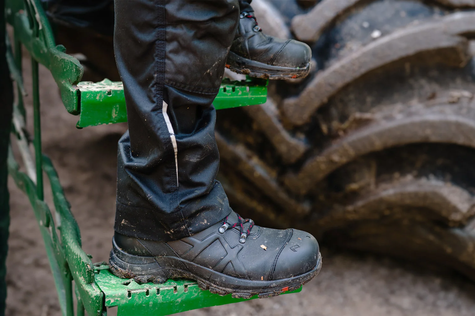 Close-up van zwarte werkschoenen met rode veters op een groene metalen trede; op de achtergrond is een grote, sterk vervuilde machineband te zien.