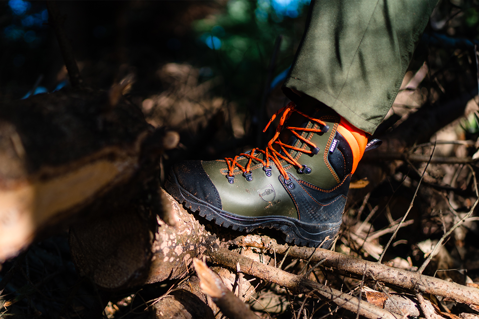 Een stevige bosbouwschoen met oranje veters staat op takken en hout in het bos, waarbij de profielzool en de hoge schacht goed zichtbaar zijn.