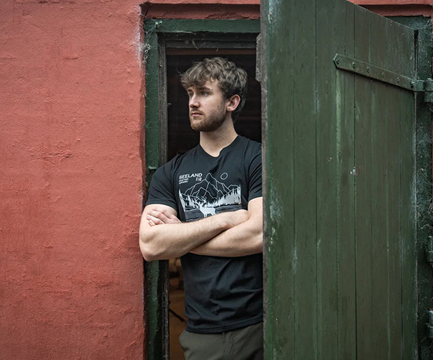 Ein junger Mann mit verschr�nkten Armen steht in einer offenen T�r und blickt nachdenklich nach drau�en.