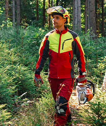 Een persoon in rood-gele bosbouwbeschermkleding en met helm loopt door het bos en draagt een kettingzaag in de linkerhand.