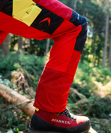 Close-up van een persoon in een rood-zwarte zaagbeschermingsbroek en werkschoen, met een gebogen been op een boomstam in het bos.