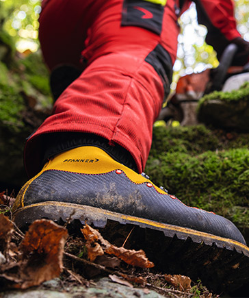 Close-up van een stevige bosbouwschoen met profielzool en gele details, gedragen door een persoon in rode beschermende kleding die zich over oneffen terrein in het bos beweegt.